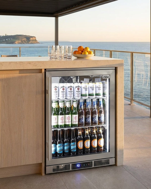 Rhino ENV 316 marine grade outdoor bar fridge built into the cabinetry of a coastal balcony bar at golden hour, ocean and headland behind